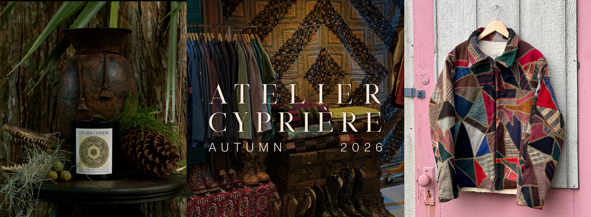 A fashion editorial banner for Atelier Cyprienne Autumn 2026, featuring a multicolor geometric patchwork jacket hanging on a pink door on the right, and a dark rustic interior with a face mask, pinecone, and patterned textiles on the left.